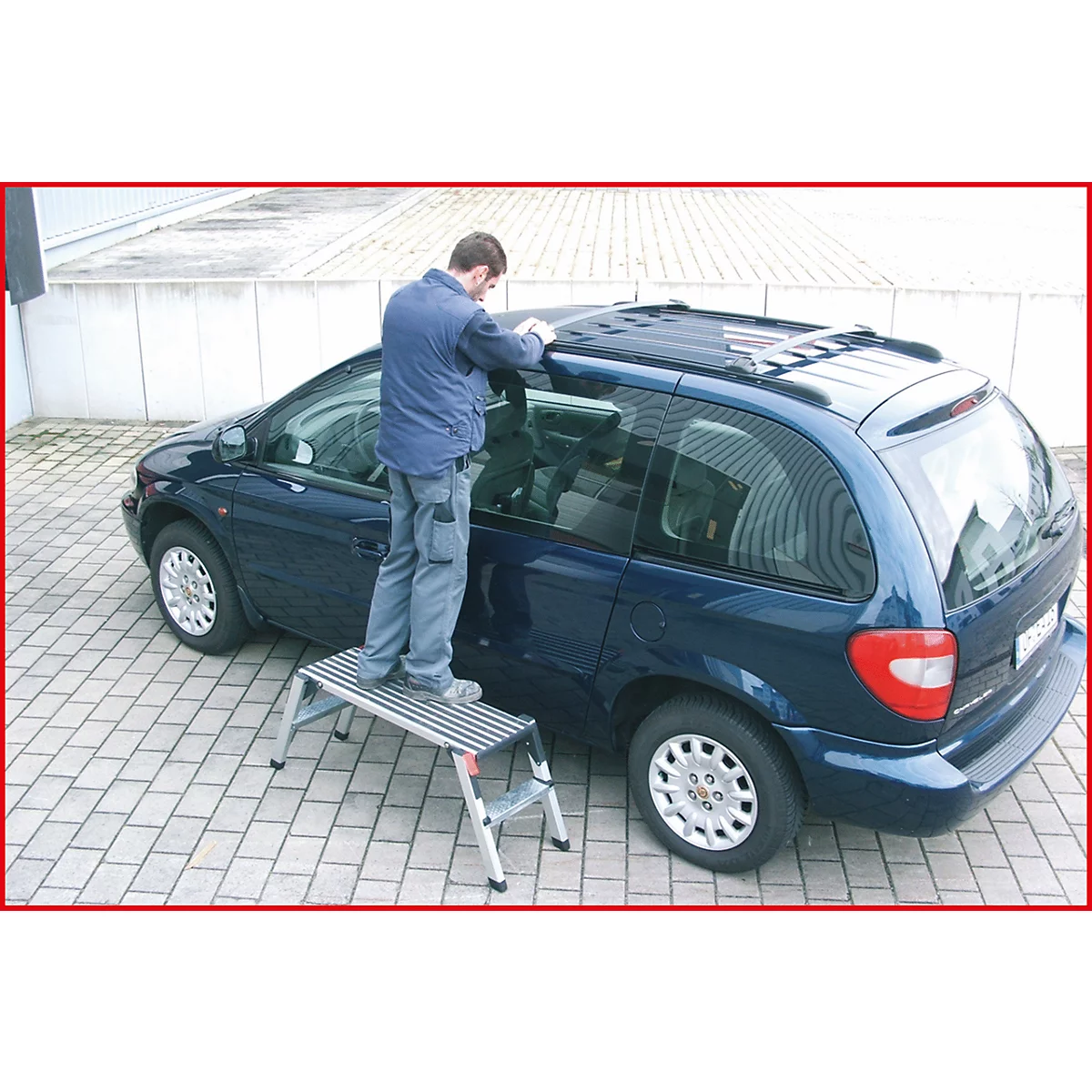 Ein Mann steht auf einer Trittleiter und arbeitet an einem blauen Van. Der Mann trägt eine Jacke und Jeans. Das Fahrzeug steht auf einer gepflasterten Fläche.